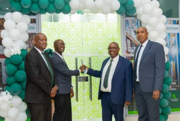 Four men in suits at a bank opening, exchanging a ceremonial key in front of a green patterned door framed by teal and white balloons.