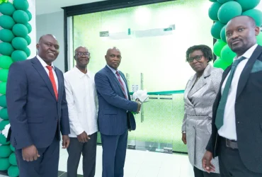 Group of five professionals in suits and dresses stand near a glass door with green geometric backdrop, celebrating a ribbon-cutting ceremony.