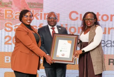 Three people on a stage; a man in a dark suit receives a framed certificate from two smiling women, one in an orange blazer and the other in a beige vest.