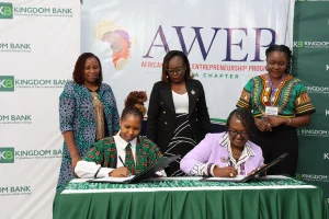 Kingdom-Bank--AWEP- Signing-Ceremony - Kingdom Bank Ltd Two women sign documents at a formal signing ceremony while three others stand nearby, with Kingdom Bank banners in the background and an AWER Africa banner visible.