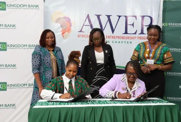 Two women sign documents at a formal signing ceremony while three others stand nearby, with Kingdom Bank banners in the background and an AWER Africa banner visible.